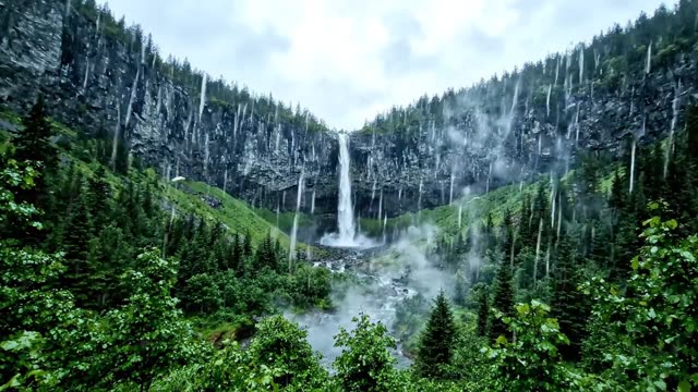 A long mountain waterfall seen across a green valley in rain
