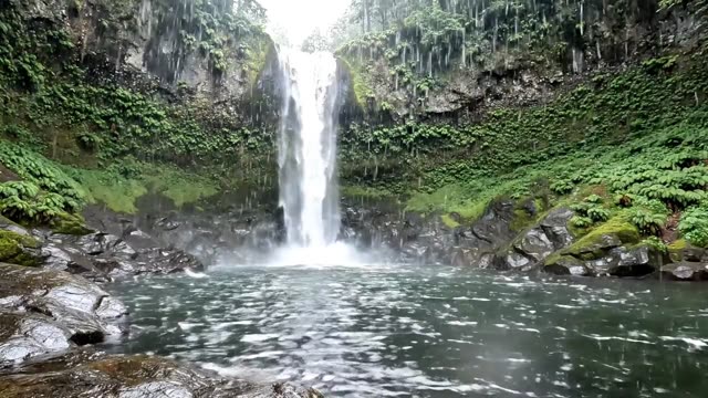 A wide waterfall dropping into a green gorge pool in rain